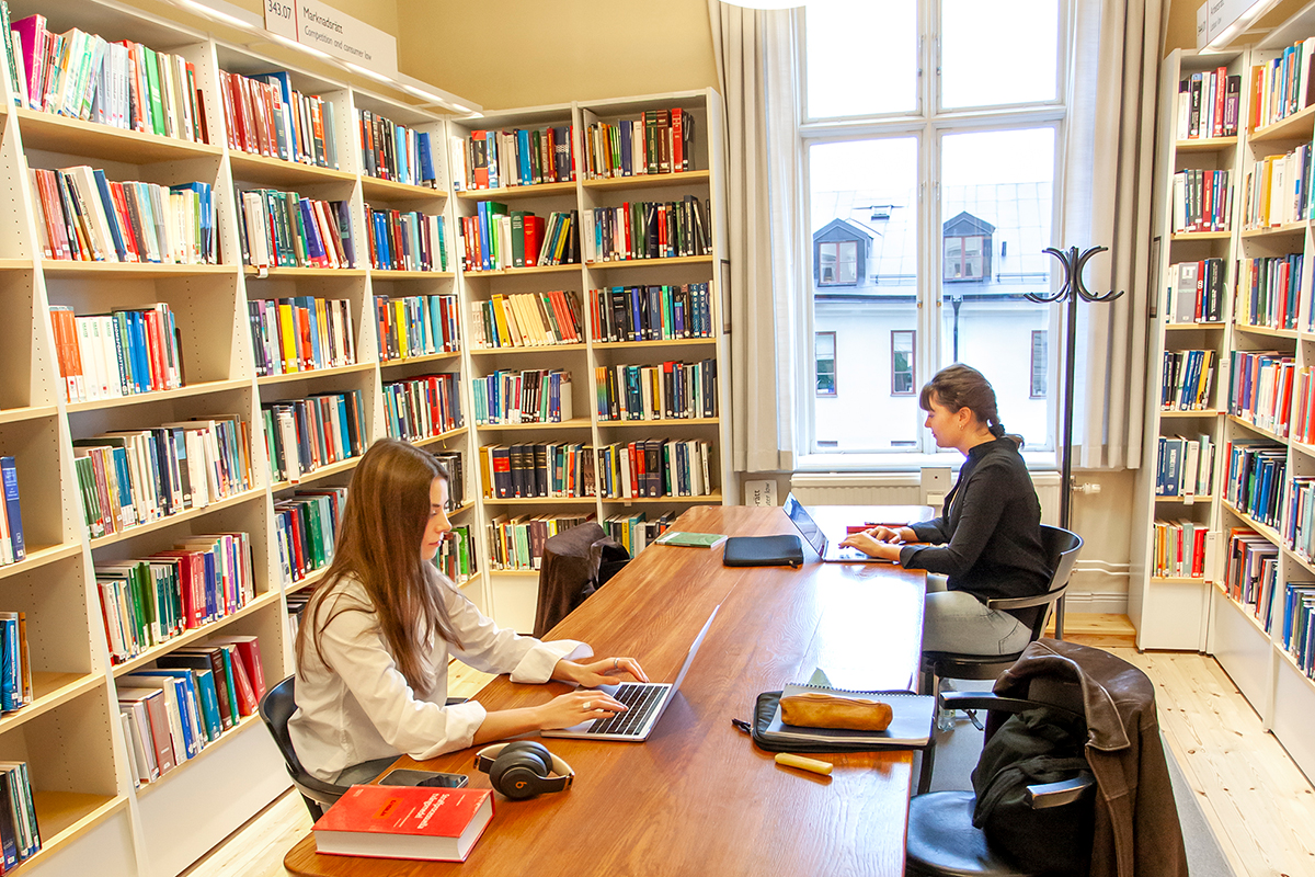 Två personer sitter och pluggar vid ett stort bord. Längs väggarna står bokhyllor fyllda av böcker.