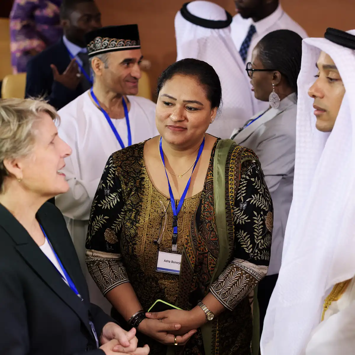People in suits and traditional dress. One person is wearing a name badge on a lanyard around their neck, as at a conference.
