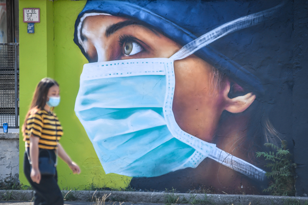 En kvinna med munskydd promenerar framför en väggmålning föreställande en kvinna med munskydd.
