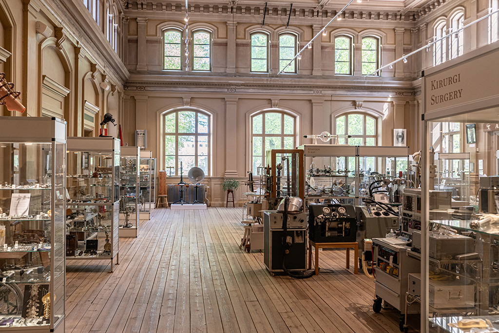 Medicinhistoriska museets stora sal. Där står glasmontrar och stora apparater. I fonden höga fönster med grönskande träd utanför.