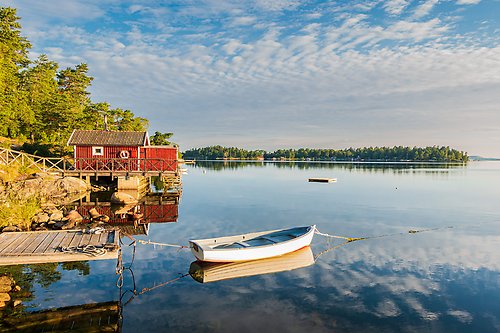 Ett soligt sommarlandskap med en röd stuga, en brygga och en roddbåt vid en spegelblank sjö.