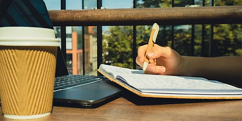Student pluggar utomhus på ett café. Studenten har kaffe, en laptop och ett skrivhäfte.