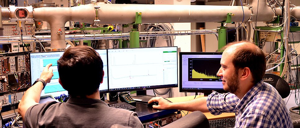 Two men in a laboratory seen from the back. They sit in front of three computer screens and seem to discuss some data shown on one screen. In the background a beamline, cables and scientific instruments are visible.
