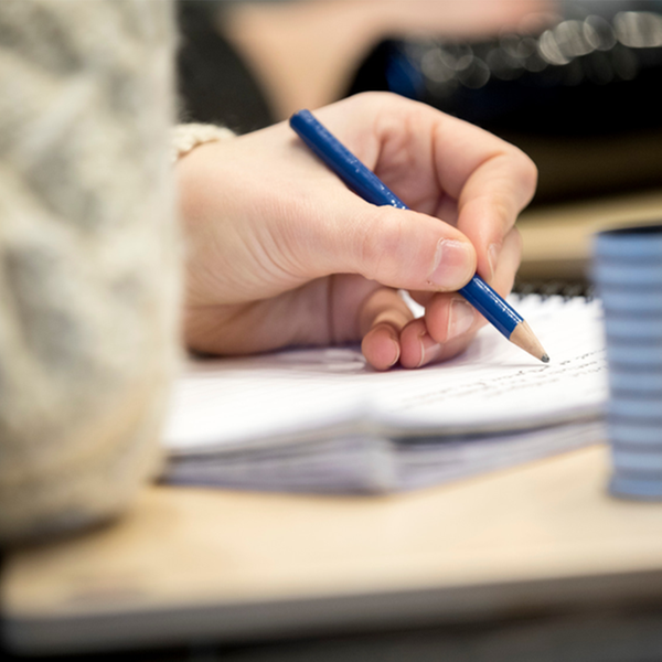 En hand som håller i en penna och skriver på ett block på en skolbänk. Foto: Mikael Wallerstedt