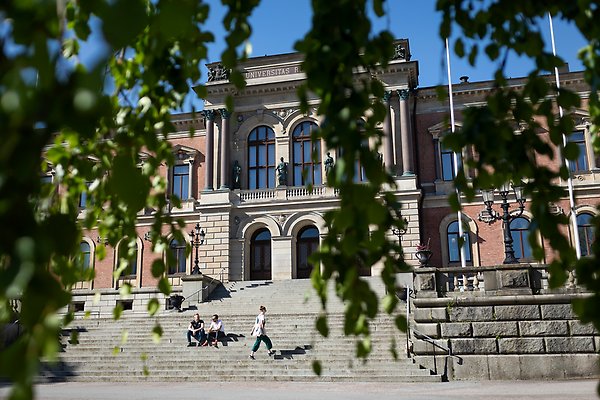 Bilden föreställer universitetshuset en sommardag. Några personer promenerar framför trappan upp till entren.