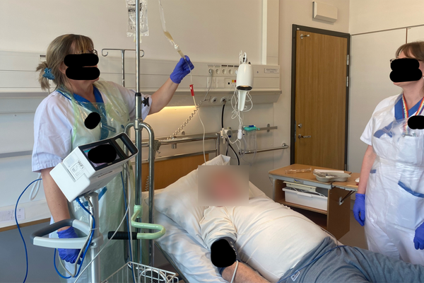 Två personer i sjukhuskläder bredvid en person i en sjukhussäng. Ansiktena är maskade.