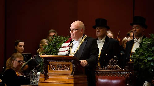 Anders Hagfeldt gives a speech at the lectern in the grand auditorium. The orchestra and the ceremonial guards stand behind him.