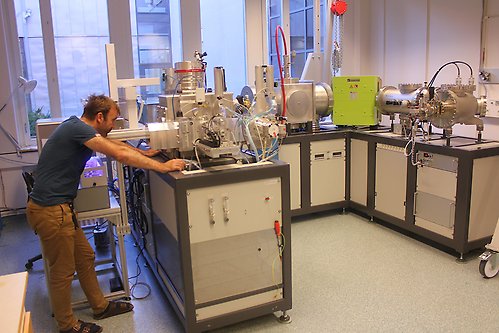 En man står bredvid en MICADAS-accelerator och ser ut att titta in i en av dess kammare. Acceleratorn är en U-formad uppställning av vakuumkammare ovanpå skåp. Dess tydligaste kännetecken är en grön magnet i ett av hörnen.