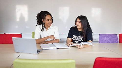 Two female students sitting by a desk and talking in a classroom
