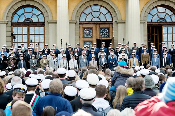 Members of male choir in suits and student caps and suits stand on the steps of Carolina Rediviva and sing in front of a crowd where many also wear student caps