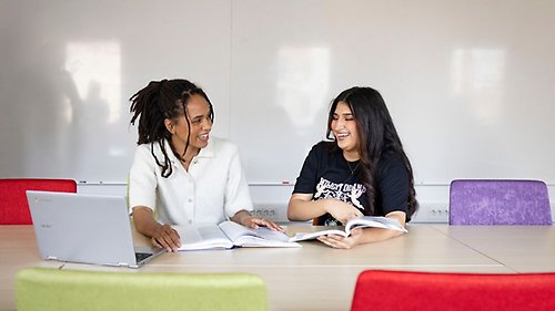 Två studenter sitter i ett seminarierum med uppslagna böcker framför sig och skrattar.