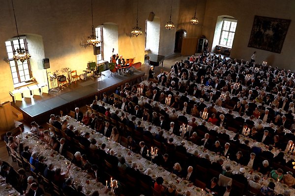Many long tables with guests in festive attire in the Hall of State at Uppsala Castle. At the side there is a stage. Chandeliers hanging from the ceiling.