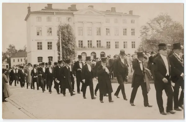 Black-and-white photograph showing a long line of men wearing top hats and black coats walking in procession up Biskopsgatan in Uppsala, with the then State Institute for Racial Biology in the background.