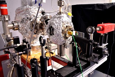 Experimentell uppställning för optiska mätningar. En ljuskälla är påslagen och lyser genom en apertur på ett prov. Bakom den optiska linjen syns två kammare av rostfritt stål som är inpackade i aluminiumfolie samt flera rör och kablar.
