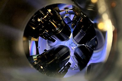 View from the top into the sputtering chamber. Four black cylinders, the magnetrons, are mounted to the chamber ceiling. Below them several pieces of material are visible. The chamber is filled by a purple plasma.
