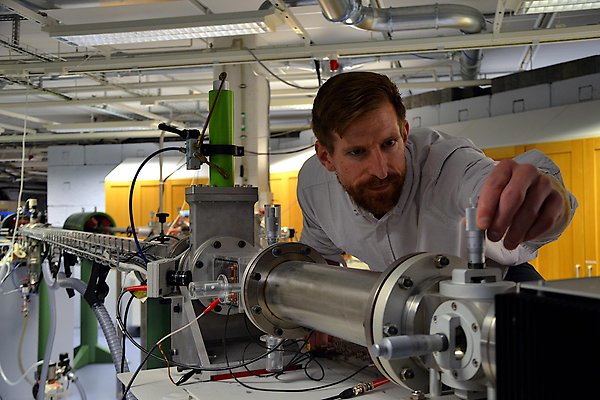 A long beamline is stretching all the way through the lower half of the photo. At the end closest to the camera, a man is leaning over the beamline to adjust a micrometer.