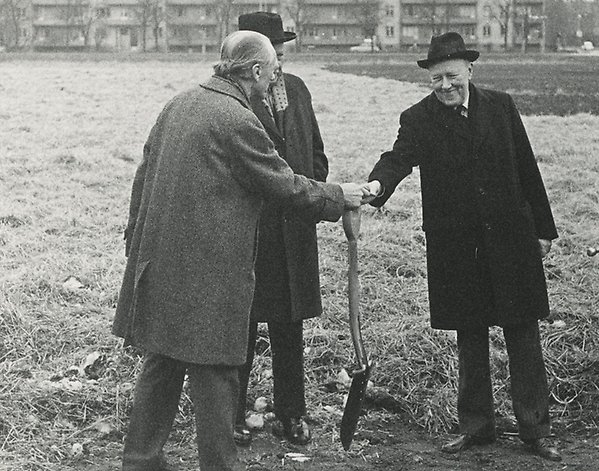 Three men in overcoats stand in a field. One of them hands a shovel to another.