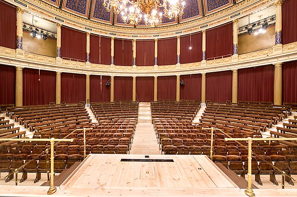 The grand auditorium seen from the center of the stage. The rows of benches stand in a semicircle. There are parquet seats right in front of the stage and around them are the terrace seats. The rows of benches are divided by five aisles, one of which is in the middle and leads from the entrance. The center of the stage has like a small balcony that sticks out and can be reached by stairs on either side. There are railings along the stairs. The balcony seats are covered with draperies.