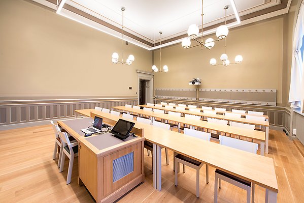 Hall I seen from the front right corner. A lectern stands at the front of the room. In front of it four rows of long tables with chairs. On the lectern there is a screen where the technology is managed. On the far wall is a projector.