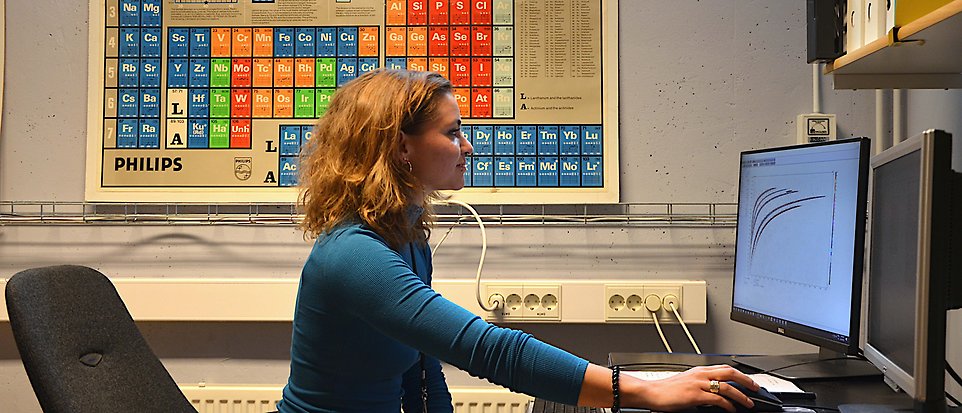 En kvinna sitter framför en datorskärm som visar vetenskapliga data. På väggen bakom henne hänger periodiska systemet.