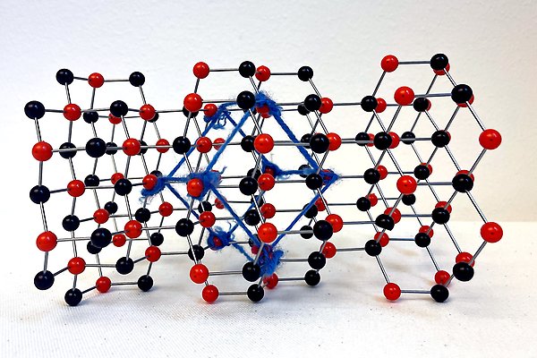 Modell av en kristall tillverkad av svarta och röda plastkulor. Ett blått snöre förbinder åtta närliggande röda kulor och bildar en kub.