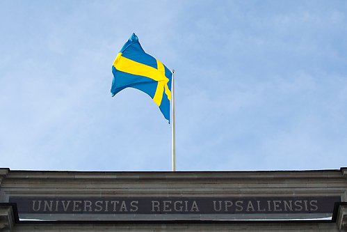 A Swedish flag, blue sky and the top of a building with the inscription Universitas Regia Upsaliensis.