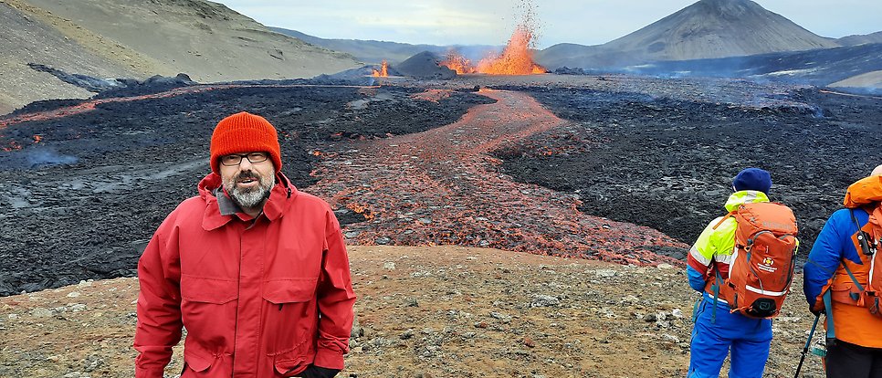 Valentin Troll in red jacket and hat in front of an erupting volcano.